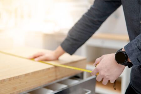Asian man interior designer using tape measure for measuring size of wooden countertop in modern kitchen showroom in furniture store. Shopping material design for home improvement.の写真素材