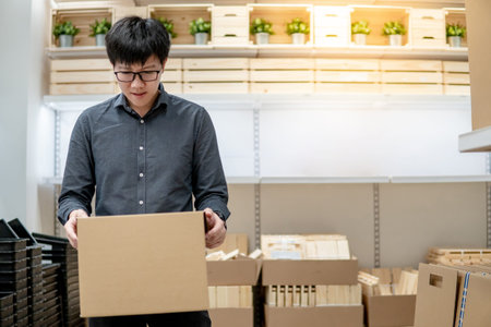 Asian courier man carrying cardboard box near product shelves in warehouse. Parcel shipping service...