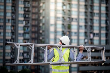 Asian maintenance worker man with safety helmet and green vest carrying aluminium step ladder at construction site. Civil engineering, Architecture builder and building service conceptsの写真素材
