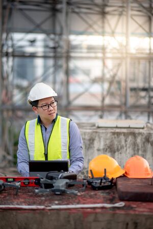 Asian engineer man working with drone, laptop and working tools at construction site. Male worker using unmanned aerial vehicle (UAV) for land and building site survey in civil engineering project.の写真素材