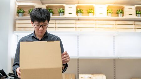 Asian courier man carrying cardboard box near product shelves in warehouse. Parcel shipping service or delivery packaging conceptの写真素材