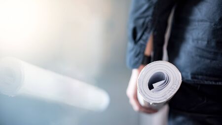 Male architect or civil engineer hand holding rolled blueprints or architectural drawing paper at construction site. Architecture and building construction industry.の写真素材