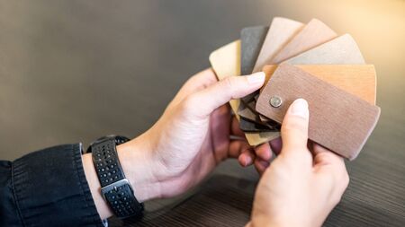 Male architect or interior designer hand holding wooden color swatch choosing wood material for housing project. Sample of wood laminated chipboard for furniture design. Architecture and constructionの写真素材