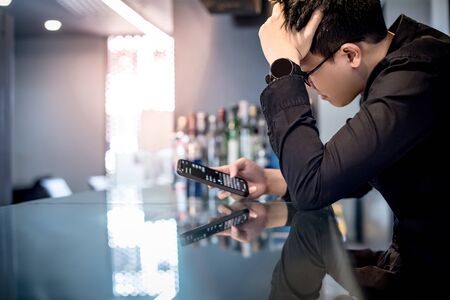 Young Asian man feeling stress, tired and headache while using chat app on smartphone at counter bar. Communication problem on social media mobile application. Cyberbullying conceptの写真素材