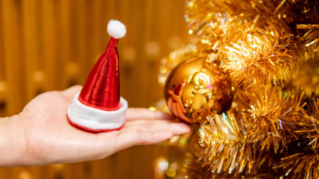 Male hand holding mini santa hat near gold Christmas tree with decorative Christmas dacoration. Xmas and New Year Celebration conceptの写真素材