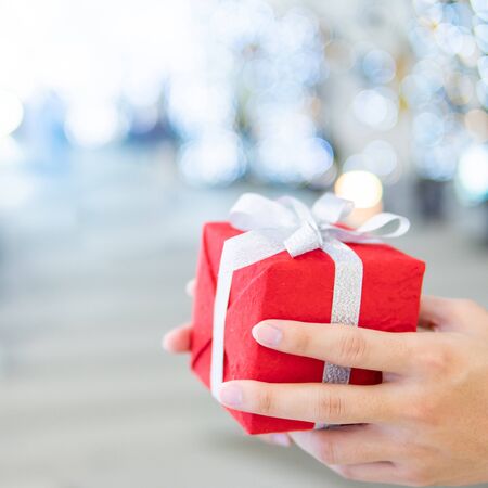 Male hand holding red Christmas gift box or New Year present with blurred illuminated decorative Christmas lights in the background. Decoration for festive season. New Year Celebration conceptの写真素材
