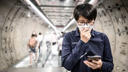 Asian man wearing surgical face mask using smartphone in subway tunnel with crowded people walking pass.の写真素材