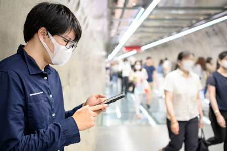 Asian man wearing surgical face mask using smartphone in subway tunnel with crowded people walking...