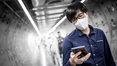 Asian man wearing surgical face mask using smartphone in subway tunnel with crowded people walking pass. coronavirus outbreak prevention in public area. Health care and medical conceptの写真素材