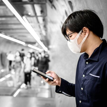 Asian man wearing surgical face mask using smartphone in subway tunnel with crowded people walking pass. coronavirus outbreak prevention in public area. Health care and medical conceptの写真素材