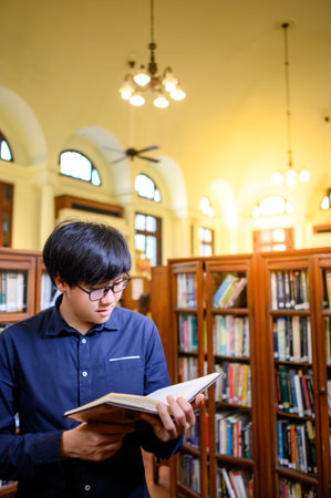 Smart Asian man university student wearing glasses reading book by vintage bookshelf. Textbook...