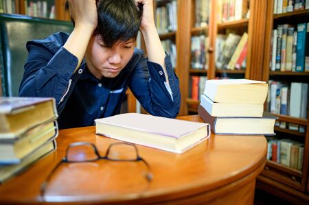Smart Asian man university student feeling stressed and anxious while reading book by vintage bookshelf in college library for education subject and research. Scholarship for education opportunity.の写真素材