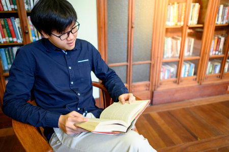 Smart Asian man university student sitting by vintage bookshelf reading book. Textbook resources in...