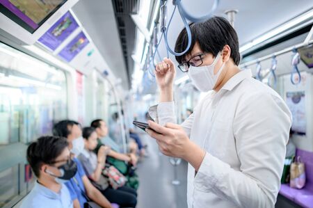 Asian man wearing surgical face mask using smartphone on skytrain or urban train. coronavirus (COVID-19) outbreak prevention in public transportation. Health awareness for pandemic protectionの写真素材