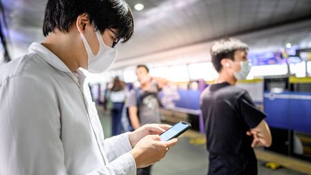 Asian man wearing surgical face mask using smartphone at subway station platform. coronavirus (COVID-19) outbreak prevention in public transportation. Health awareness for pandemic protectionの写真素材