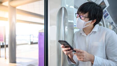 Asian man wearing surgical face mask using smartphone on urban skytrain or subway. coronavirus (COVID-19) outbreak prevention in public transportation. Health awareness for pandemic protectionの写真素材