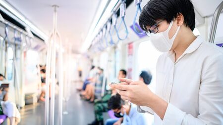 Asian man wearing surgical face mask using smartphone on skytrain or urban train. coronavirus (COVID-19) outbreak prevention in public transportation. Health awareness for pandemic protectionの写真素材