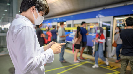 Asian man wearing surgical face mask using smartphone at skytrain station platform. coronavirus...
