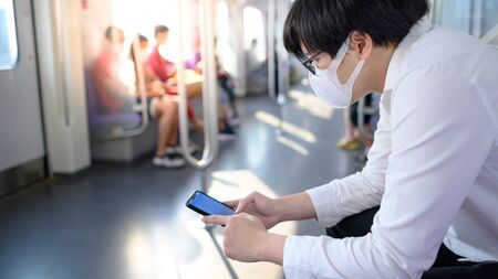 Asian man wearing surgical face mask using smartphone on skytrain or urban train. coronavirus (COVID-19) outbreak prevention in public transportation. Health awareness for pandemic protectionの写真素材