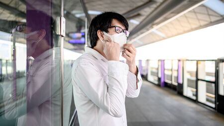 Asian man wearing face mask at skytrain station or urban train platform. coronavirus (COVID-19) outbreak prevention in public transportation. Health awareness for PM2.5 air pollution protection.の写真素材