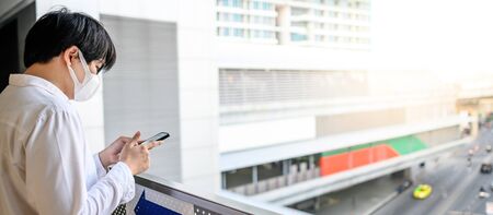 Asian man wearing surgical face mask using smartphone at skytrain station platform.  coronavirus (COVID-19) outbreak prevention in public transportation. Health awareness for pandemic protectionの写真素材