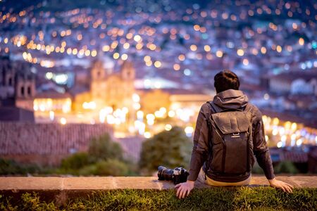 Asian man traveler and photograpaher sitting on viewpoint looking at illuminated cusco city at night. Cusco (Cuzco) is a city in southeastern Peru, near the Urubamba Valley of the Andes mountain rangeの写真素材