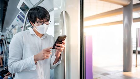 Asian man wearing surgical face mask using smartphone on urban skytrain or subway. coronavirus (COVID-19) outbreak prevention in public transportation. Health awareness for pandemic protectionの写真素材
