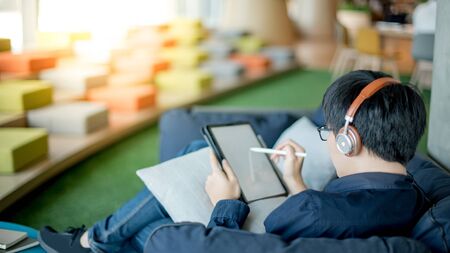Asian businessman with headphones listening to music working with digital tablet on comfortable sofa at home office. Man with glasses doing online conference on mobile app. Work from home conceptの写真素材