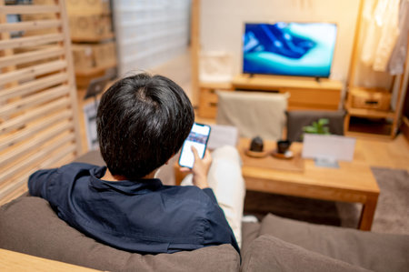 Young Asian man in casual clothing sitting on comfortable sofa (couch) using smartphone and watching...
