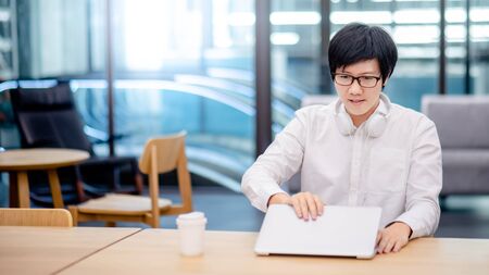 Asian man entrepreneur wearing glasses and headphones using laptop at home office. Businessman working with personal computer thinking about business project. Work smart with modern technology.の写真素材