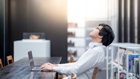 Asian man entrepreneur wearing glasses and headphones using laptop at home office. Businessman working with personal computer thinking about business project. Work smart with modern technology.の写真素材