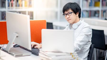 Young Asian man university student using computer and laptop for educational research in college library. Male researcher finding information on internet platform. Online study or E-learning conceptの写真素材