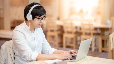Young Asian man wearing headphones listening to music while working with laptop computer. Male office worker doing online conference with his business company team. Work from home conceptの写真素材