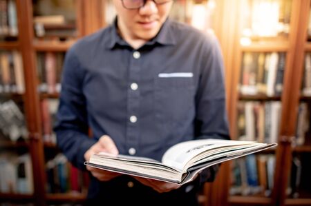 Smart Asian man university student wearing glasses reading book by vintage bookshelf. Textbook resources in college library for educational subject and research. Scholarship for education opportunity.の写真素材
