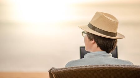 Asian man sitting on beach chair with his laptop computer at tropical island beach looking at the sea when sunset. Summer holiday or vacation travel conceptsの写真素材