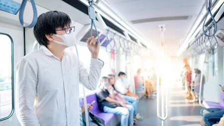 Asian man wearing surgical face mask holding handrail on skytrain or urban train. Coronavirus (COVID-19) outbreak prevention in public transportation. Health awareness for pandemic protectionの写真素材