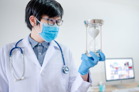 Healthcare and medical procedure process concept. Male Asian doctor or physician man wearing surgical mask and latex gloves holding silver hourglass in exam room at hospital clinic. の写真素材