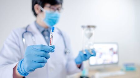 Male Doctor or physician man holding COVID-19 test tube and silver hourglass in hand with blue latex gloves. Coronavirus vaccine waiting period conceptの写真素材