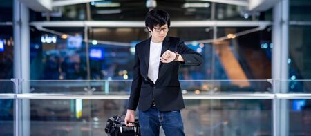 Business travel and baggage claim check-in concept. Smart Asian businessman wearing formal suit jacket carrying suitcase luggage and backpack waiting for the flight in international airport terminal.の写真素材