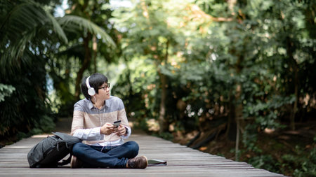 Young Asian man with headphones, glasses and backpack sitting on wooden bridge relaxing in the park....