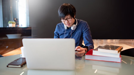 Smart Asian business man with glasses and headphones using laptop computer while reading book in...
