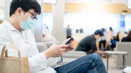 Male Asian patient wearing surgical face mask using smartphone at waiting area in hospital or medical center. Medical exam or body check up. Coronavirus pandemic prevention. Health care conceptの写真素材