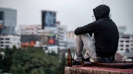 Mystery man in white mask with hoodie jacket sitting with broken alcoholic drink bottle on abandoned building feeling tired and stressed. Depression, mental health problem or alcoholism conceptの写真素材