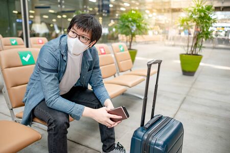 Asian man tourist wearing protective face mask sitting with suitcase luggage in airport terminal. Coronavirus (COVID-19) pandemic prevention when travel abroad. Health awareness and social distancingの写真素材