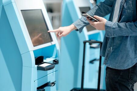 Male tourist holding passport and smartphone using self check-in kiosk in airport terminal. Travel abroad conceptの写真素材