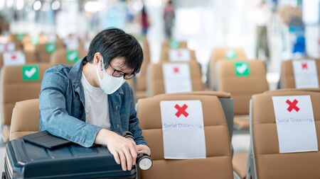 Asian man tourist wearing face mask opening suitcase luggage while sitting in airport terminal. Coronavirus (COVID-19) pandemic prevention when travel abroad. Health awareness and social distancingの写真素材