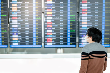Asian man tourist wearing face mask holding suitcase luggage at arrival departure board in airport...