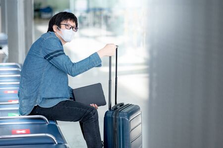 Asian man tourist wearing face mask holding digital tablet and suitcase luggage in airport terminal. Coronavirus (COVID-19) prevention when travel abroad. Health awareness and social distancingの写真素材