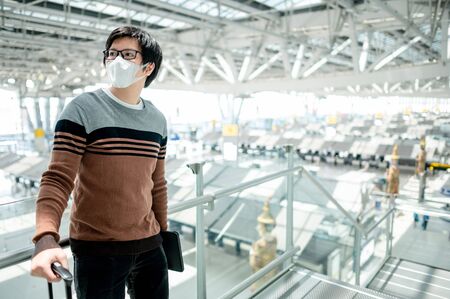 Asian man tourist wearing face mask carrying suitcase luggage waiting for check-in in airport terminal. Coronavirus (COVID-19) pandemic prevention when travel. Health awareness and social distancingの写真素材