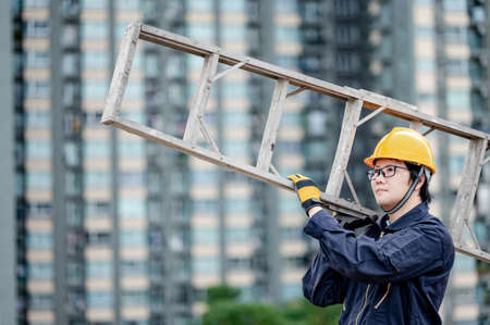 Asian maintenance worker man with protective suit and safety helmet carrying aluminium step ladder at construction site. Civil engineering, Architecture builder and building service conceptsの写真素材